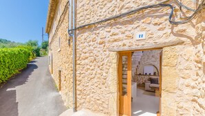 A stone building with a number '12' on the wall, a door leading to a room with a sofa and a table, and a narrow street with greenery on one side. - CASA VELLA (VALL PETIT) - Chalet with terrace in Mancor De La Vall. Free WiFi (Mancor de la Vall)