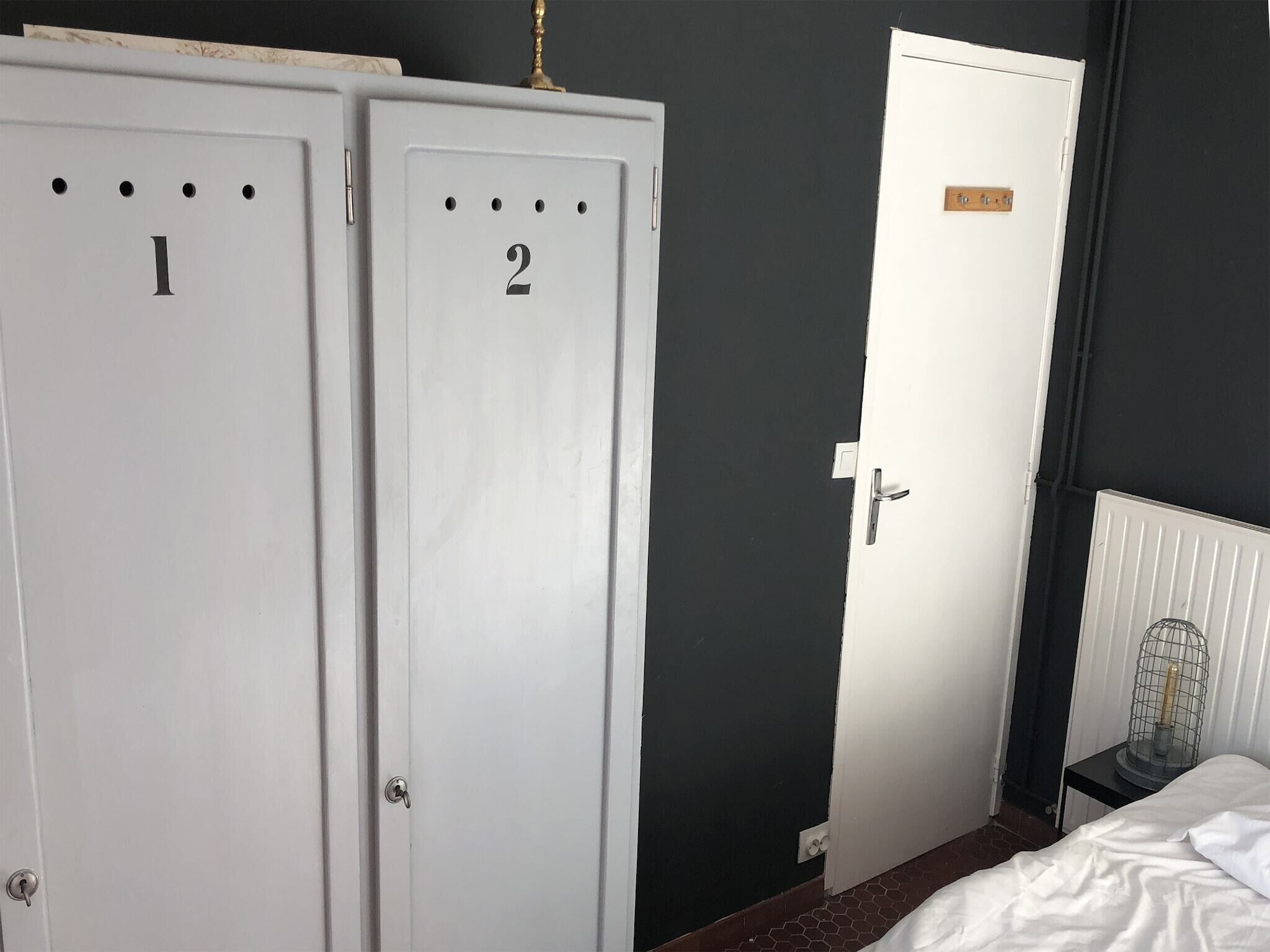 Quarto com dois armários brancos identificados com os números 1 e 2, uma cama com roupa de cama branca, um radiador e uma pequena mesa com um objeto parecido com uma gaiola.
