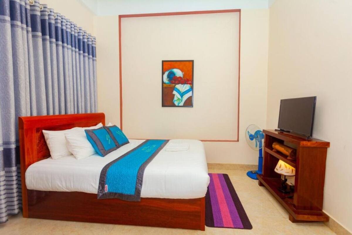 A hotel room with a bed, a television, a fan, and a painting on the wall.