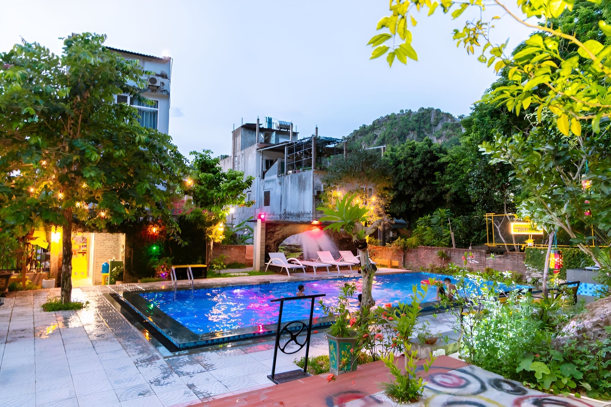 Una piscina con sillas de descanso, rodeada de follaje y un edificio con fuente de agua.