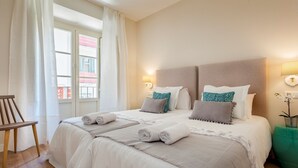 Ein ordentlich bezogenes Bett mit Kissen und Decke, ein Holzhocker sowie ein Nachttisch mit Lampe und Pflanze.