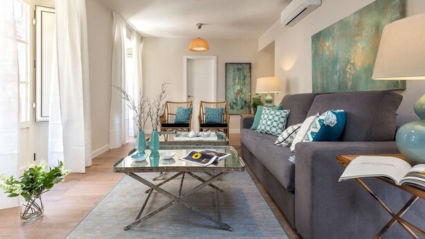 Ein modernes Wohnzimmer mit einem grauen Sofa, einem Couchtisch und einem Bücherregal. Ein Fenster mit weißen Vorhängen, ein Bild an der Wand und eine Lampe auf dem Beistelltisch sind ebenfalls zu sehen.