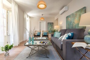 Un salon moderne avec un canapé gris, une table basse et une bibliothèque. Il y a une fenêtre avec des rideaux blancs, un tableau au mur et une lampe sur la table d’appoint.