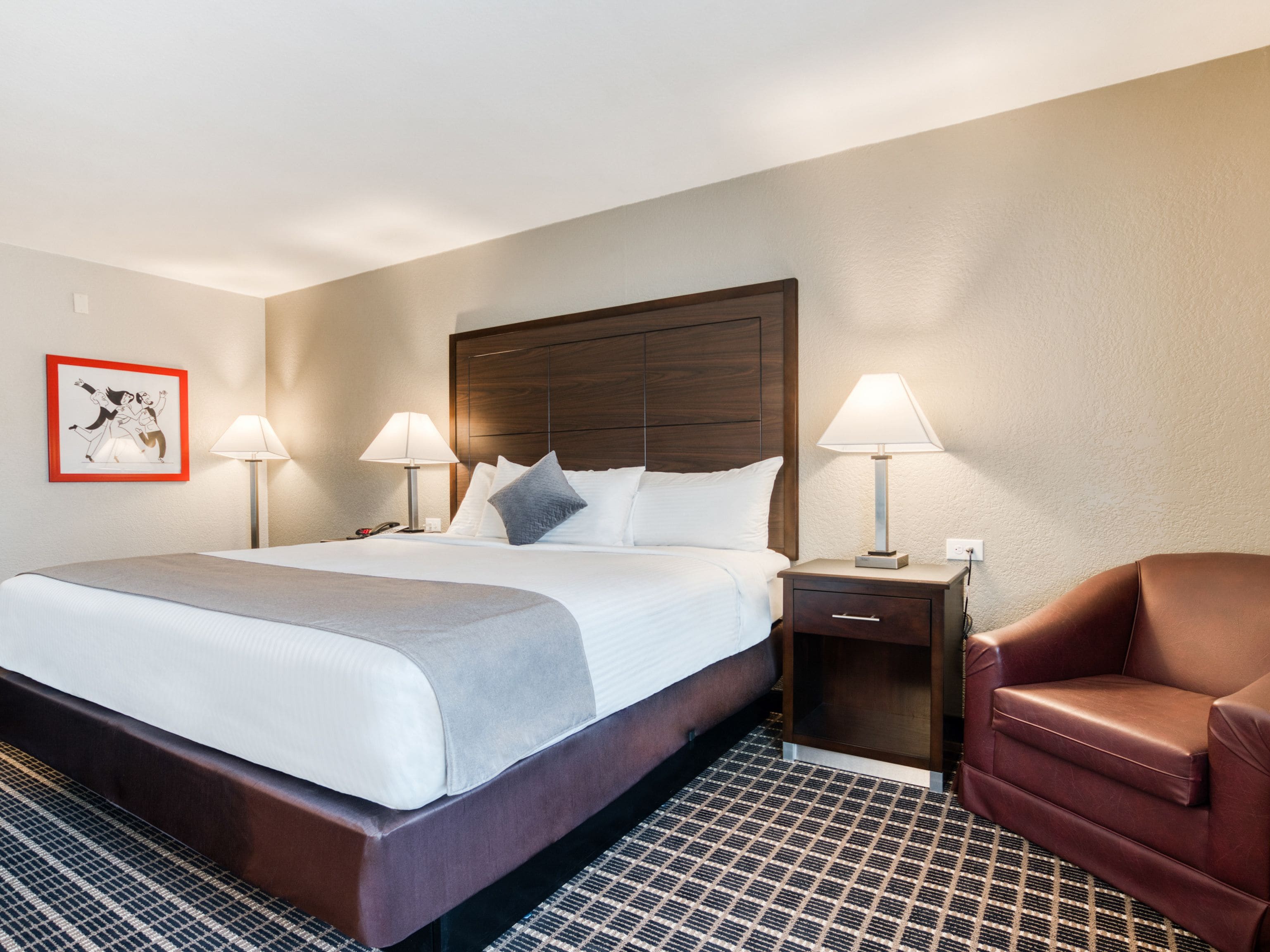 Une chambre d’hôtel avec un grand lit, deux tables de chevet avec des lampes, un fauteuil en cuir marron et une moquette à motifs.