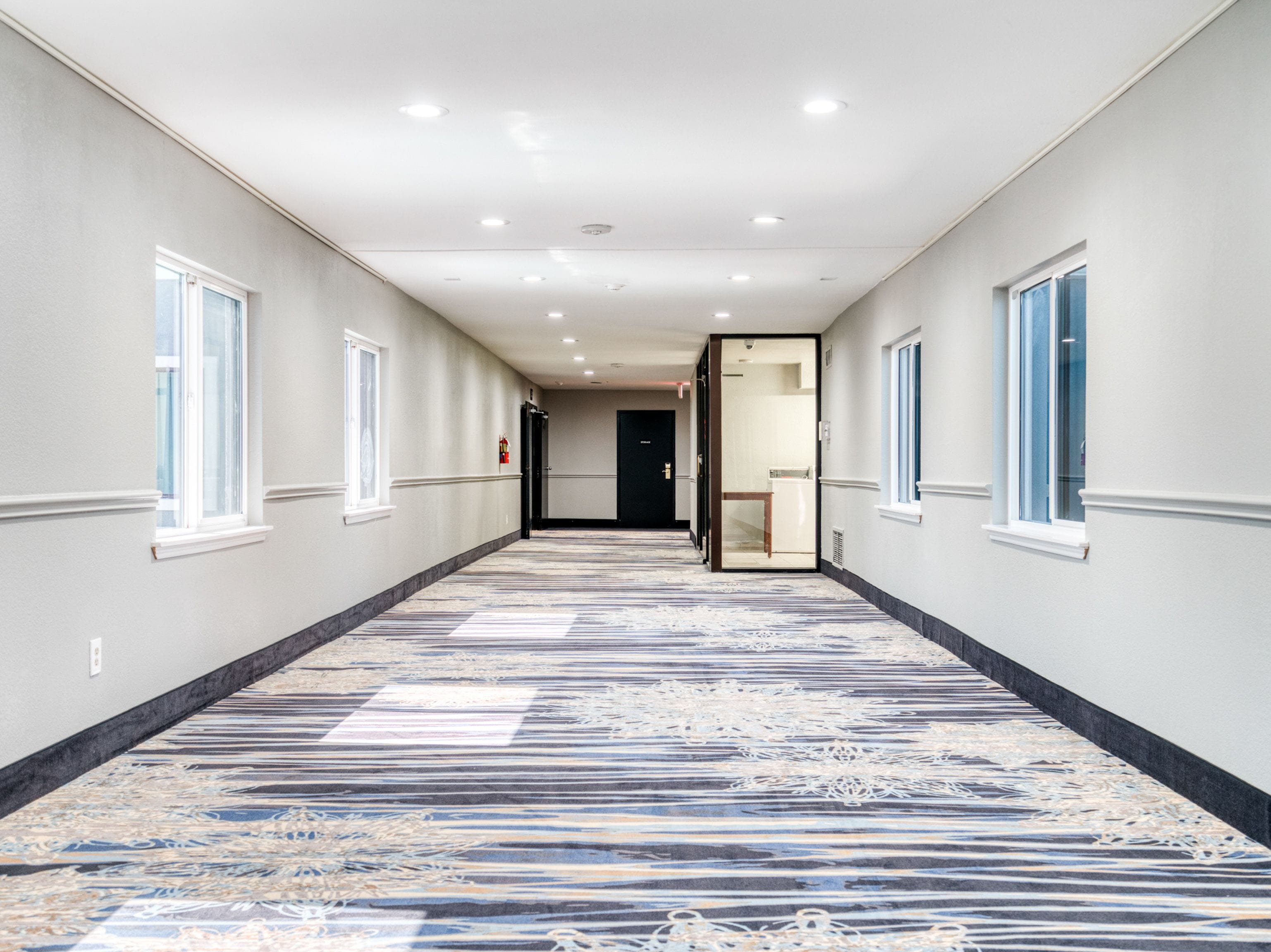 A hotel corridor with a patterned carpet, white walls, and multiple windows.