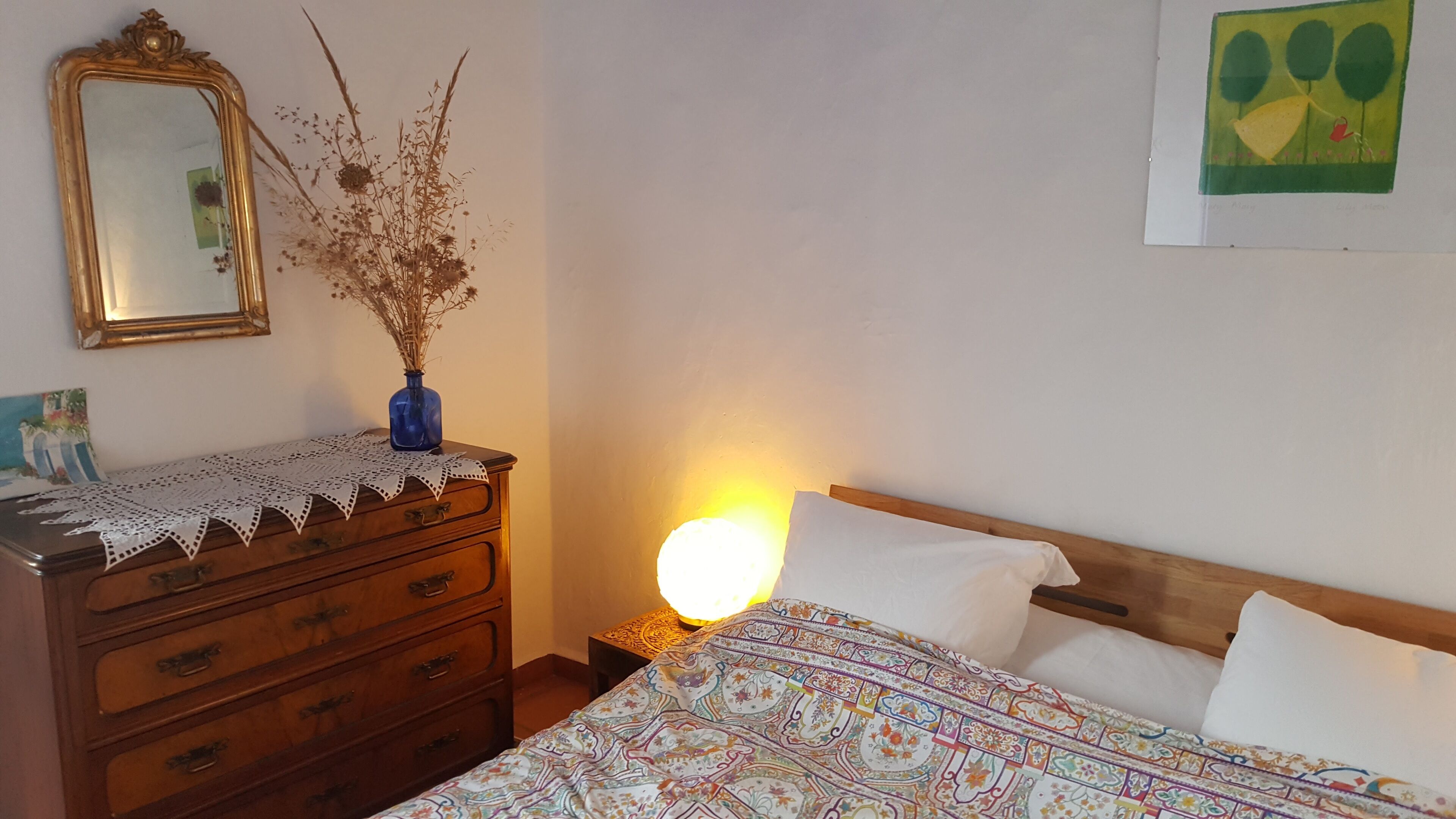 A bedroom with a wooden dresser, a mirror, a vase with dried plants, a bed with patterned bedding, and a wall painting.
