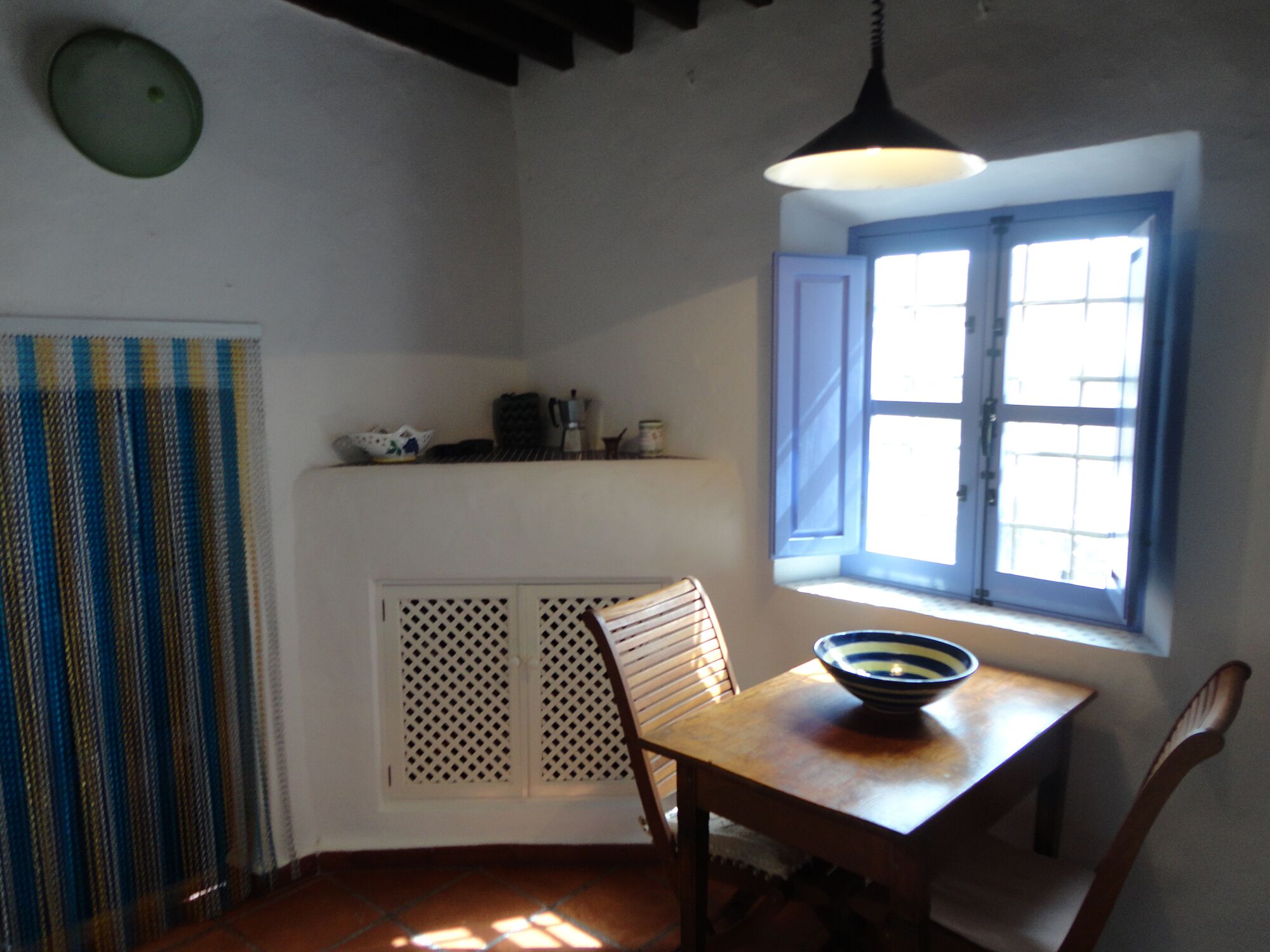 A dining area with a table, chairs, a bowl, a window with blinds, and a patterned curtain.