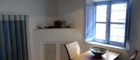 A dining area with a table, chairs, a bowl, a window with blinds, and a patterned curtain.