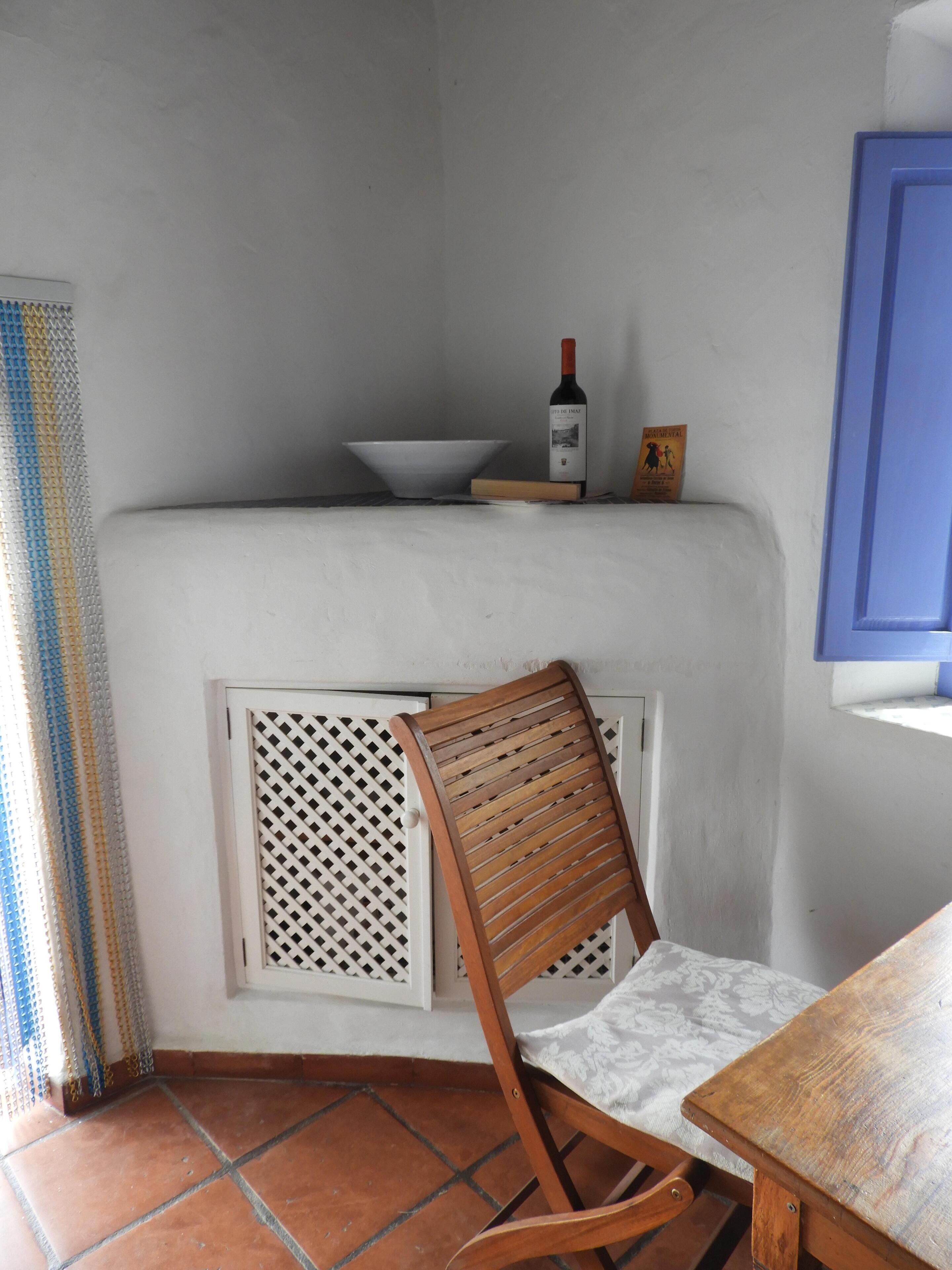 Esquina de una habitación con pared blanca, una silla de madera, una mesa, un radiador blanco y una botella de vino en un estante.