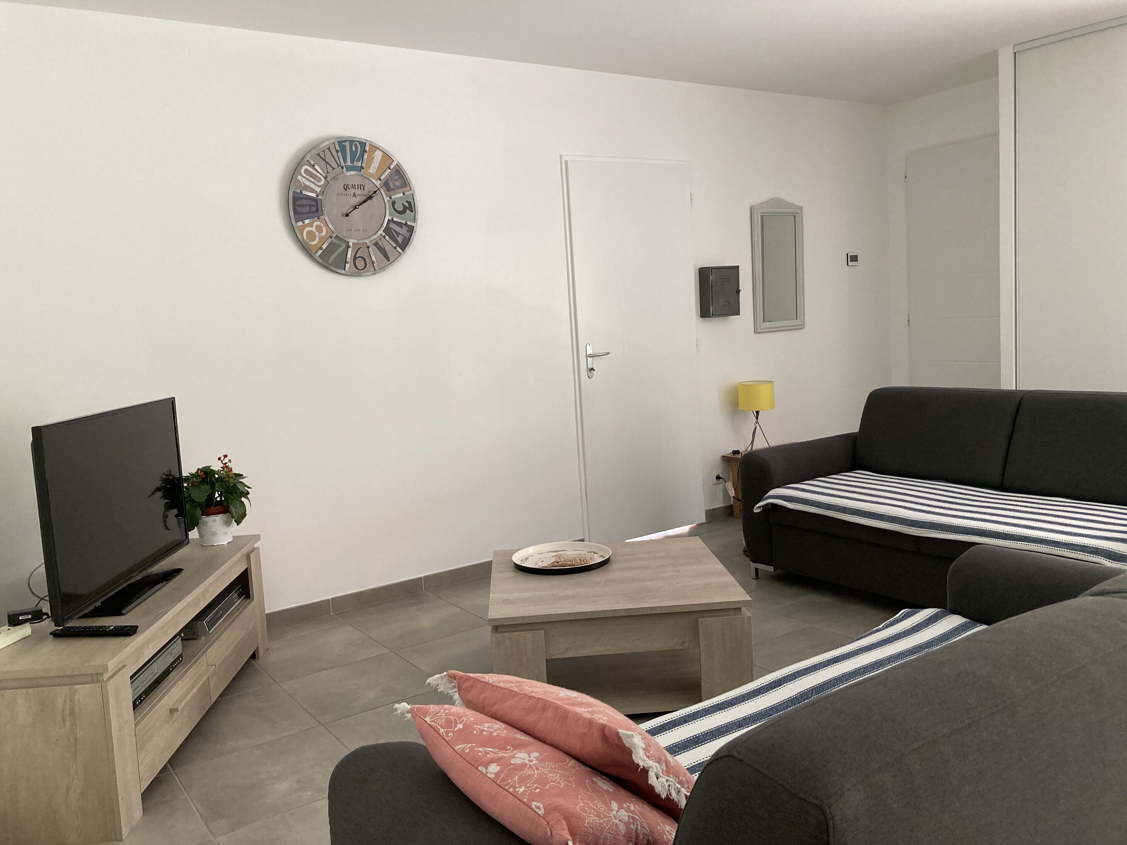 A living room with a flat-screen TV, a coffee table, a sofa, a clock, a door, and a small table with a lamp.
