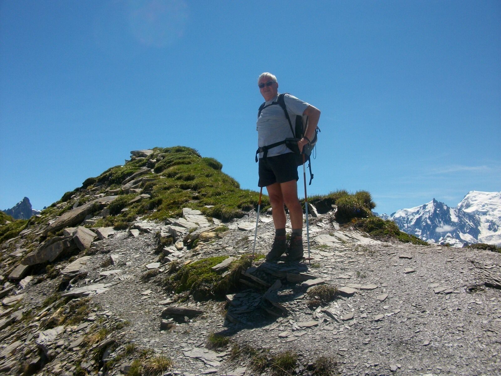 Une personne se tient sur un sentier de montagne rocheux, un sac à dos sur le dos et des bâtons de randonnée à la main, entourée de montagnes enneigées et sous un ciel bleu dégagé.
