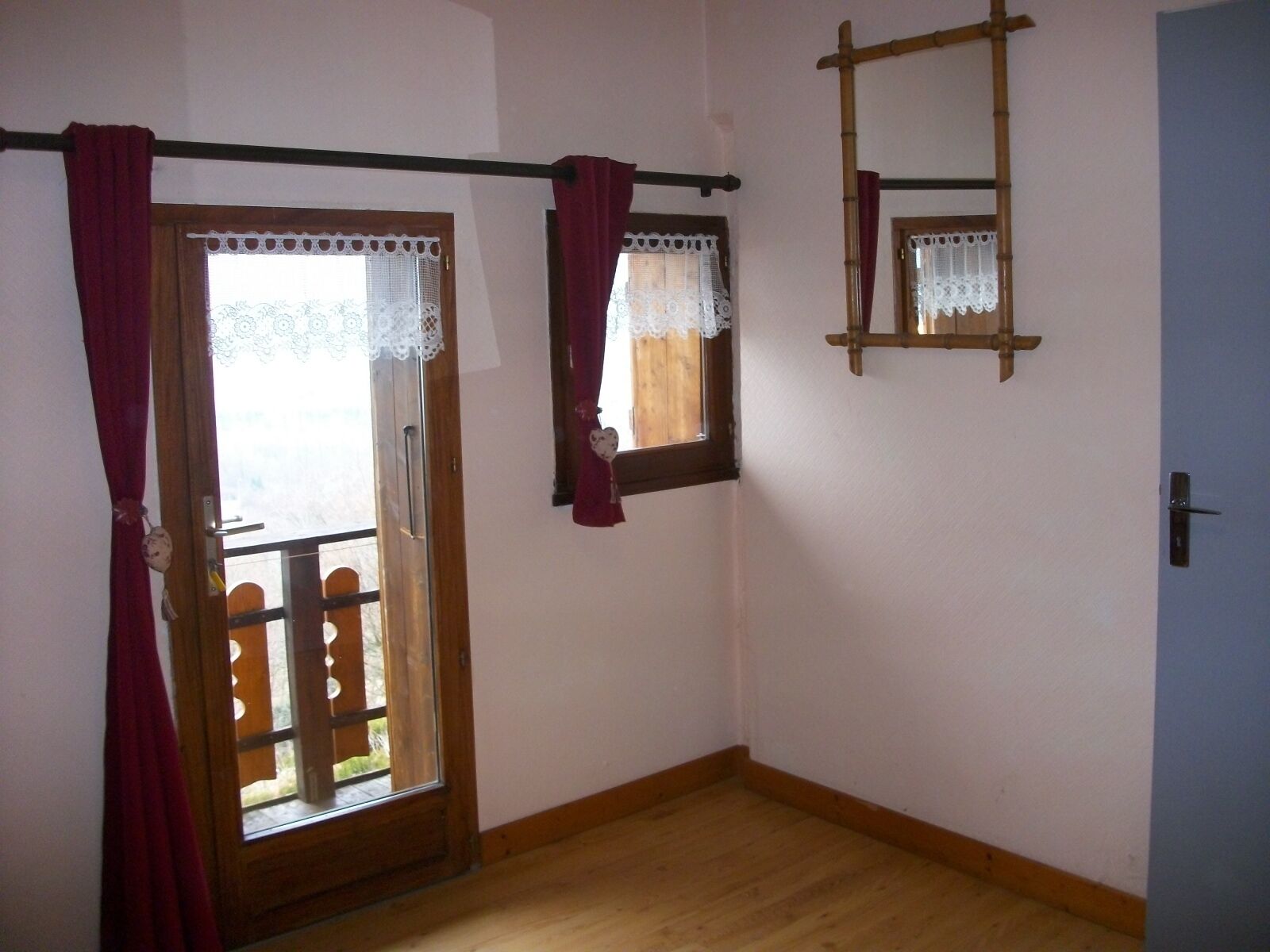 Una habitación con suelo de madera, una puerta con panel de vidrio y una ventana con marco de madera y cortinas de encaje.