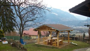 A wooden gazebo with benches and a table, surrounded by a grassy yard and a tree.