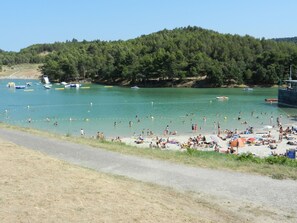 A beach with people swimming and sunbathing, boats on the water, and a forested hill in the background.