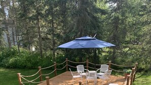 Una terraza de madera con sillas blancas y un paraguas azul, rodeada por una barandilla de cuerda y follaje.