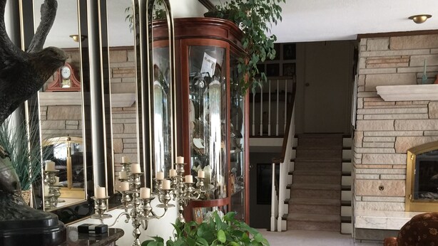 Un salón espacioso con un amplio aparador de madera, una escalera y varios objetos decorativos, incluyendo un espejo grande, una estatua y plantas en macetas.