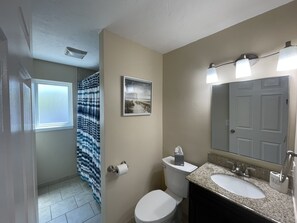 Un baño con inodoro, lavamanos y ducha. Hay un cuadro en la pared y un perchero para toallas.