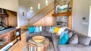A spacious living room with a gray sectional sofa, wooden coffee table, and a stone fireplace. A wooden staircase with a railing leads to an upper level. - Amazing Ocean Views in Yachats! Modern Decor! Dog Friendly! (Yachats)