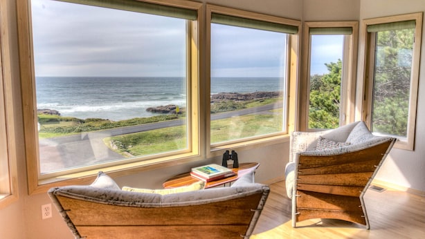A room with large windows overlooking the ocean, two wooden chairs, a small table with books, and a pair of binoculars. - Amazing Ocean Views in Yachats! Modern Decor! Dog Friendly! (Yachats)