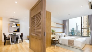 A modern hotel room with a wooden floor, a bed with white bedding, a desk with a chair, a large window with a city view, and a wooden partition.