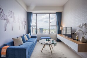 Ein modernes Wohnzimmer mit einem blauen Sofa, einem runden Couchtisch, einem Fernseher auf einer Holzkonsole und Blick auf die Stadt durch große Fenster.