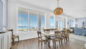 Un comedor amplio con una mesa grande y sillas, ventanas grandes con persianas y vista a la playa.