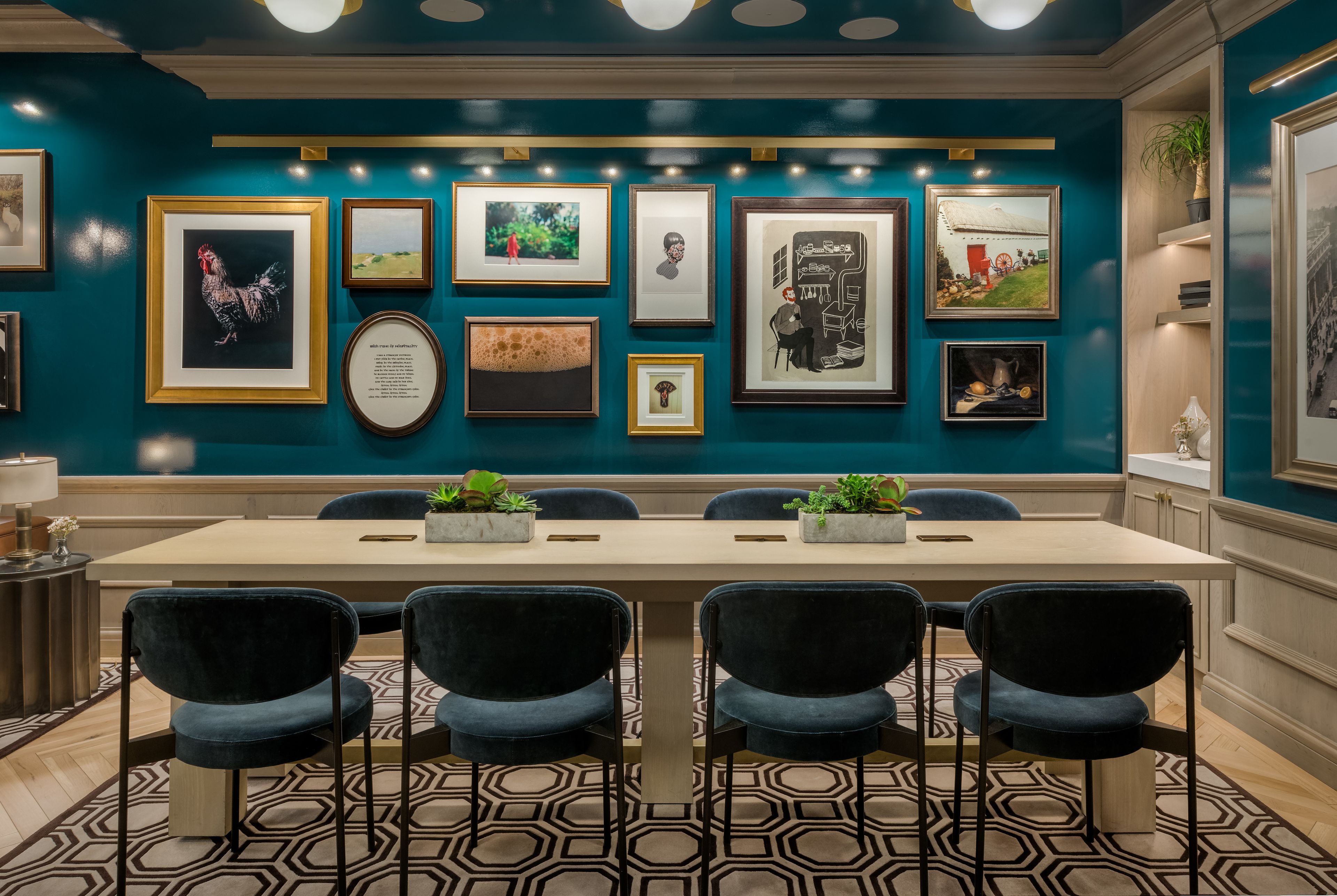 A conference room with a long table and four chairs, framed artwork on the walls, and a potted plant on the right side.