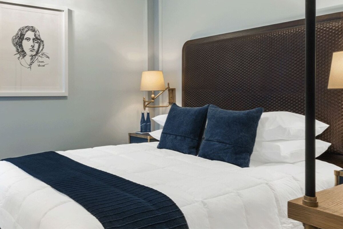 A neatly made bed with a white and navy blue bedspread, two navy blue pillows, and a navy blue throw blanket. There is a framed artwork on the wall above the bed, a bedside table with a lamp, and a nightstand with a bottle and a book.