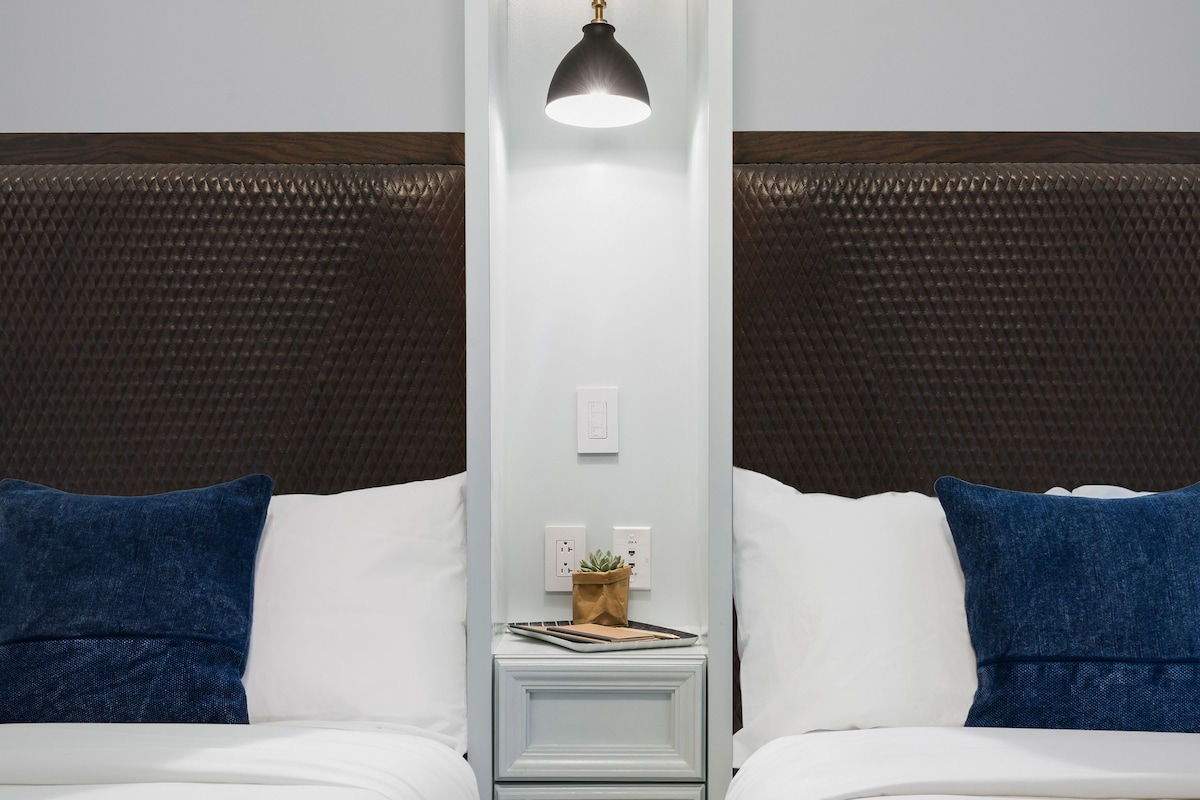 A hotel room with two beds, white bedding, a nightstand with a lamp, and a wall-mounted outlet.