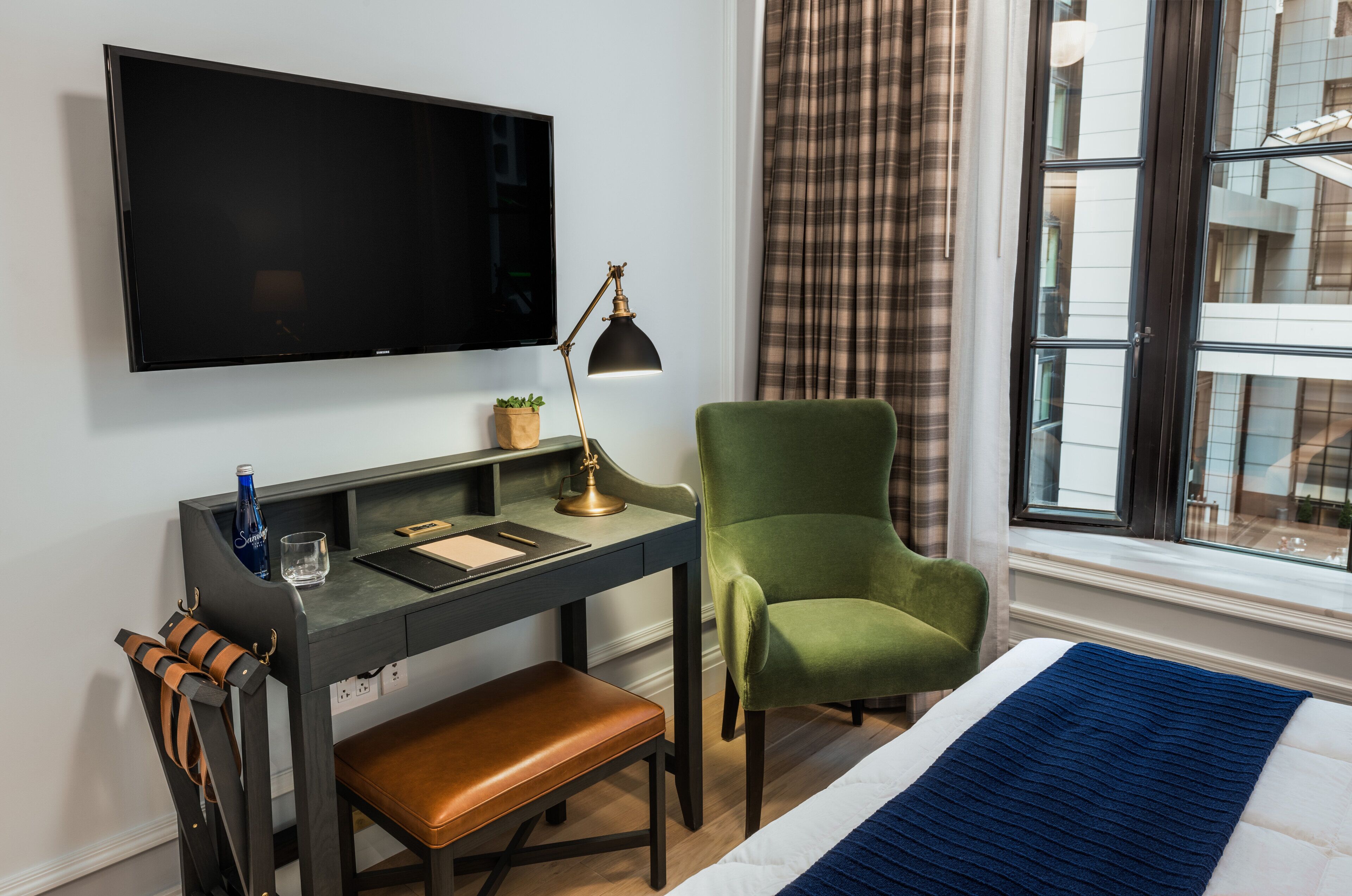 A hotel room with a flat-screen TV mounted on the wall, a desk with a lamp, a green chair, a bench, and a view of the city through a window with curtains.