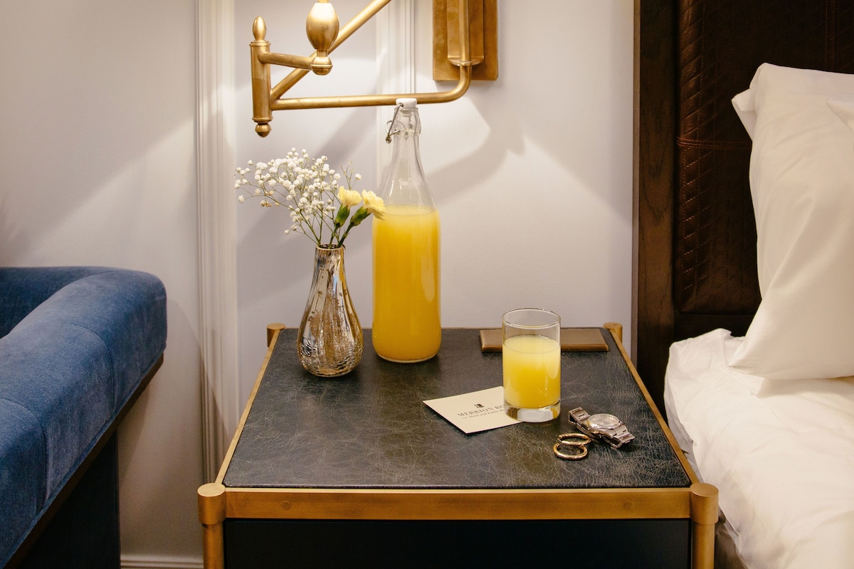 A neatly made hotel room with a bed, a bedside table with a lamp, a vase with flowers, and a bottle of orange juice.