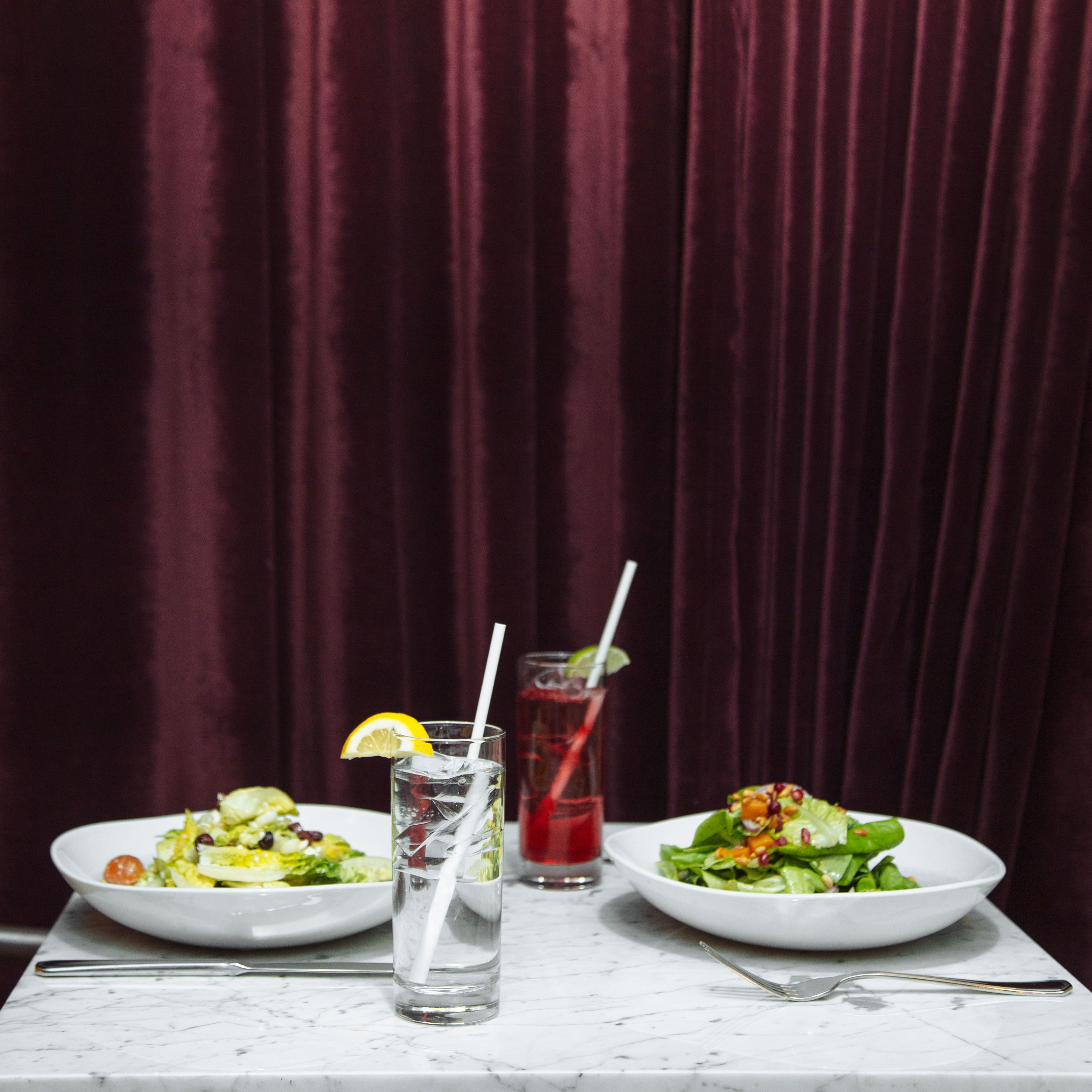 Et bord med to salatportioner på hvide tallerkener og et glas vand med sugerør.