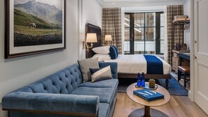Una habitació d'hotel amb un llit gran, un sofà blau, una taula petita amb un llibre i una pintura d'un paisatge de muntanya.