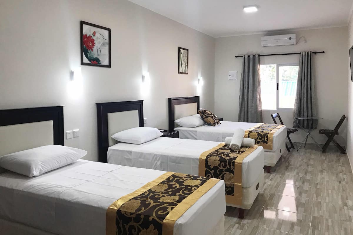 Une chambre d’hôtel avec trois lits simples, une petite table, deux chaises, une fenêtre avec des rideaux et un appareil de climatisation.