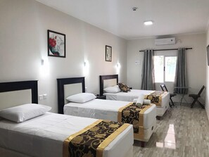 Habitación de hotel con tres camas individuales, una mesita pequeña, dos sillas, un ventanal con cortinas y un equipo de aire acondicionado.