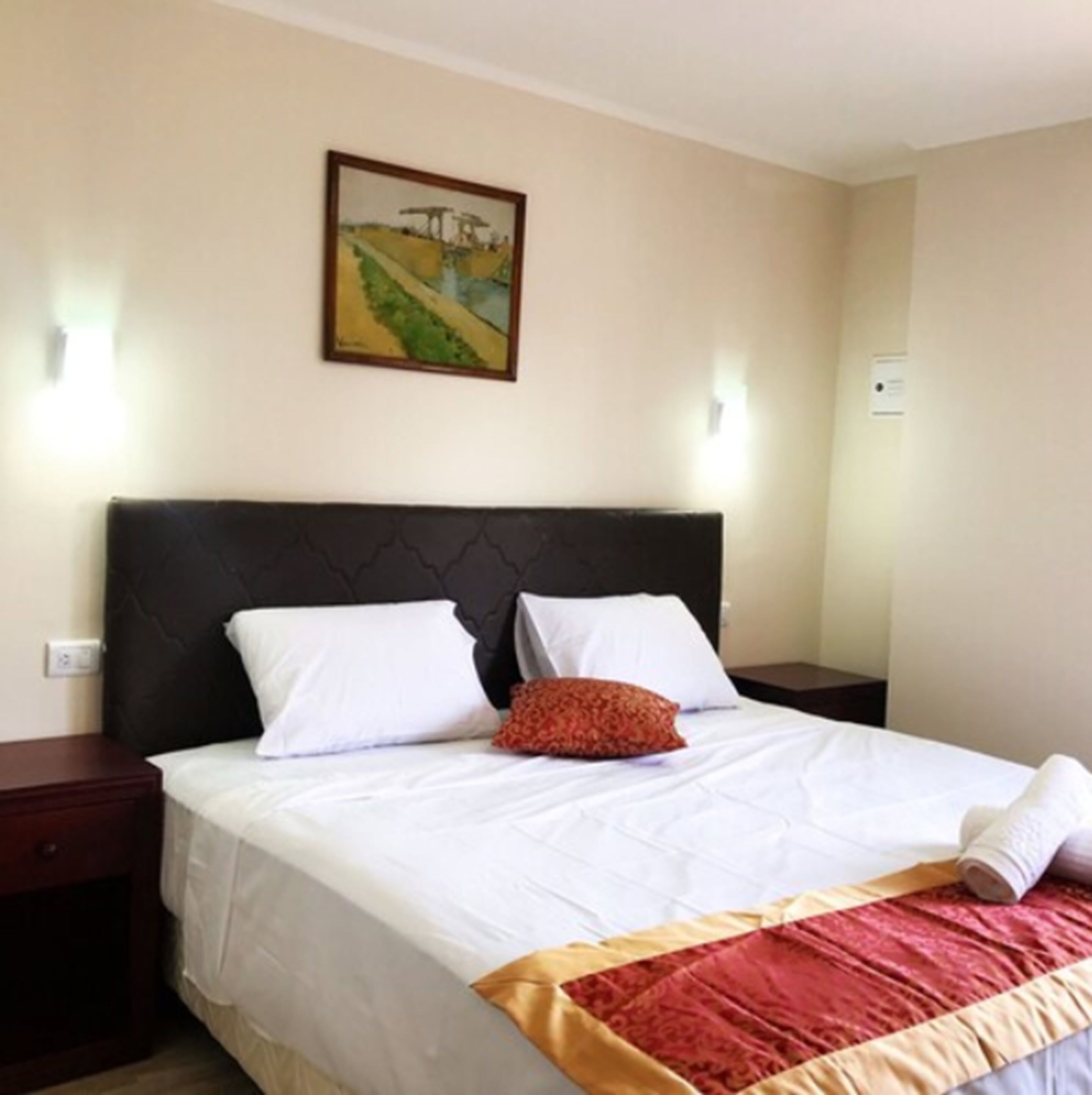 A neatly made bed with white linens and a red accent pillow, a dark headboard, and a framed painting on the wall.