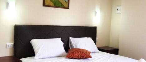 A neatly made bed with white linens and a red accent pillow, a dark headboard, and a framed painting on the wall.