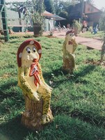 Two wooden sculptures of women in traditional attire, one holding a red ribbon, set on a grassy area with a building and trees in the background.