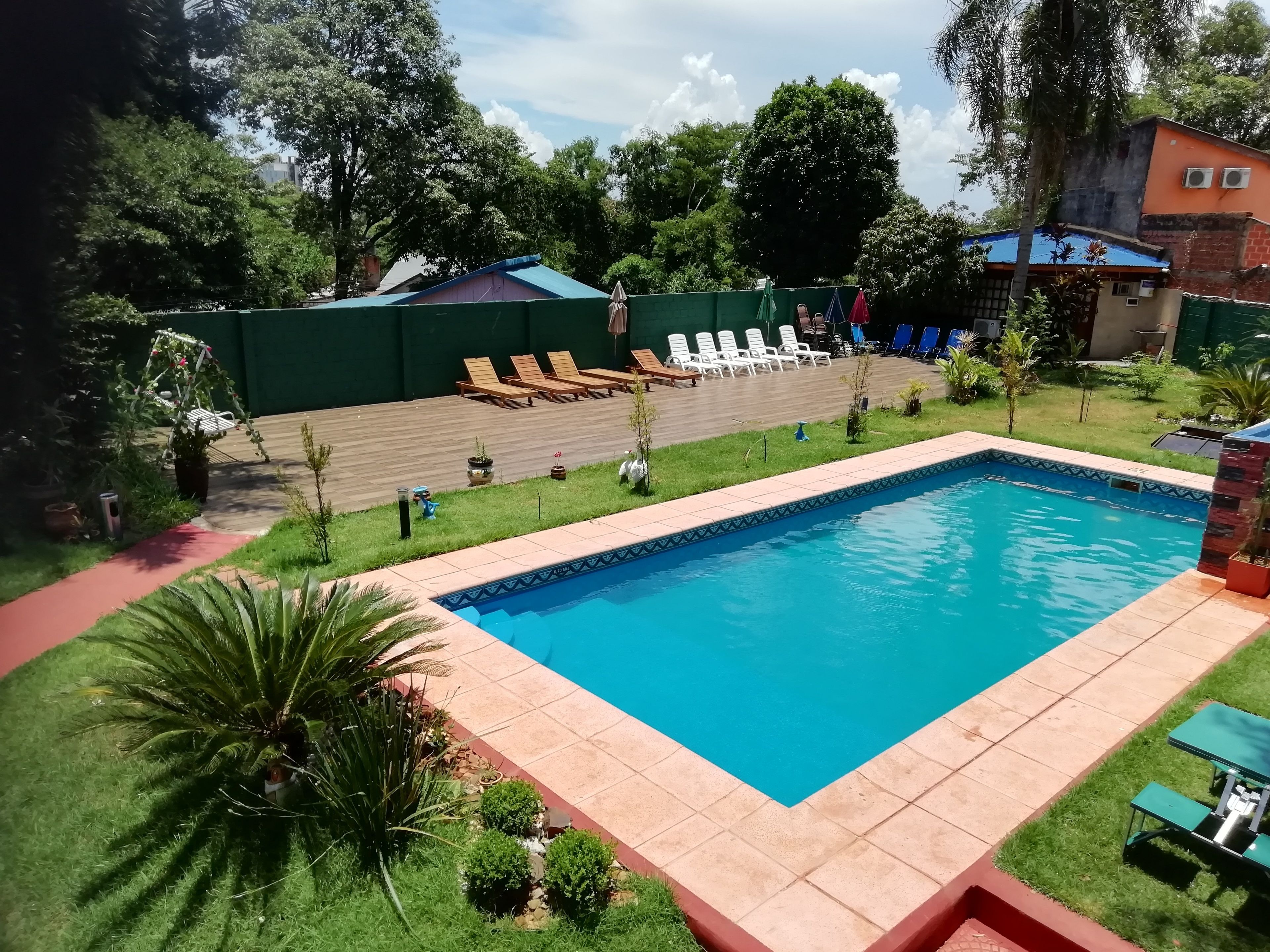 A rectangular swimming pool with a tiled deck, surrounded by green grass and small shrubs. There are lounge chairs and a small table on the deck.
