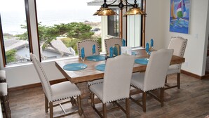 Un coin repas avec une table en bois, des chaises à revêtement blanc et une vue sur l’océan grâce à de grandes fenêtres.