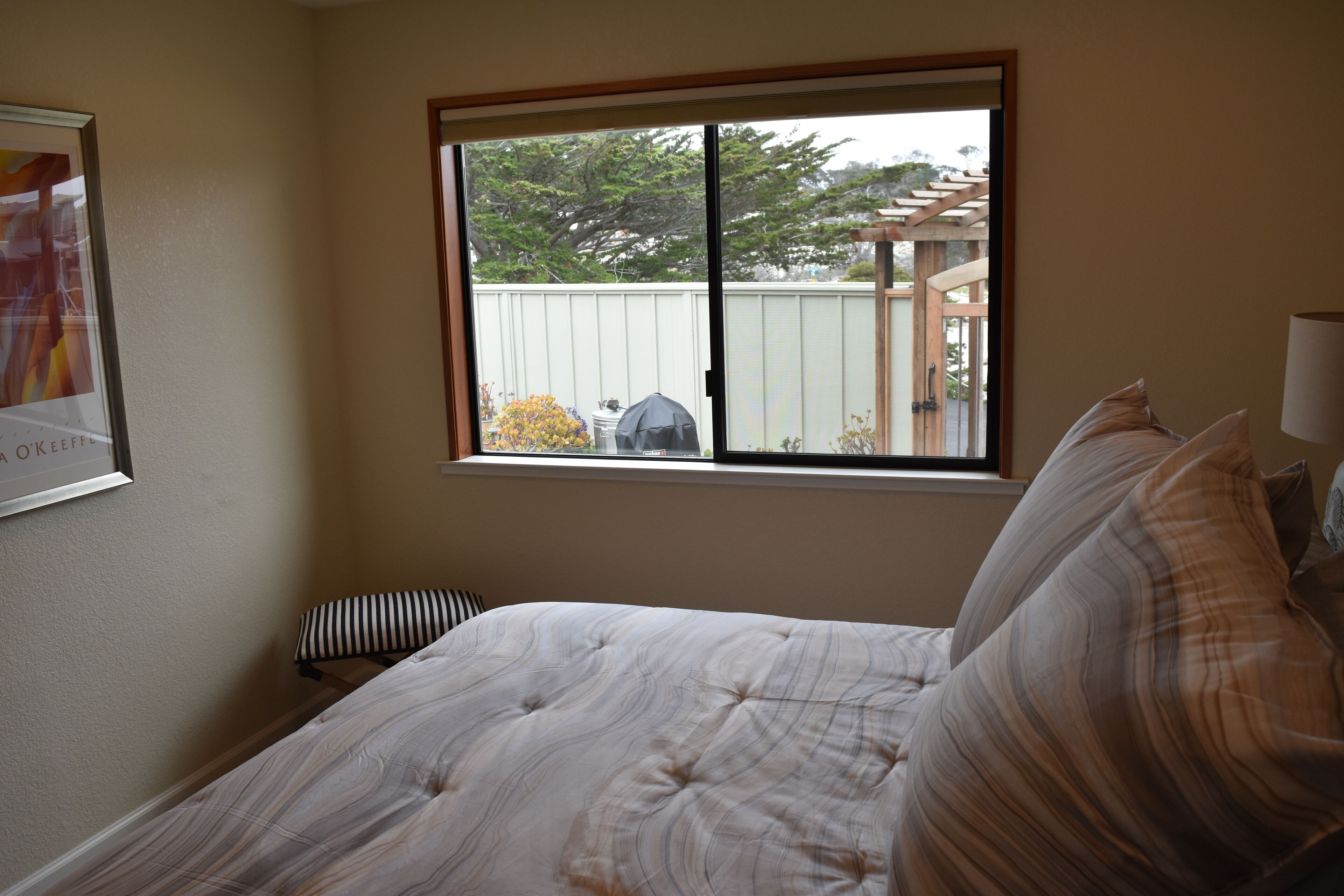A bedroom with a large window, a bed with striped bedding, a small bench, and a framed picture on the wall.