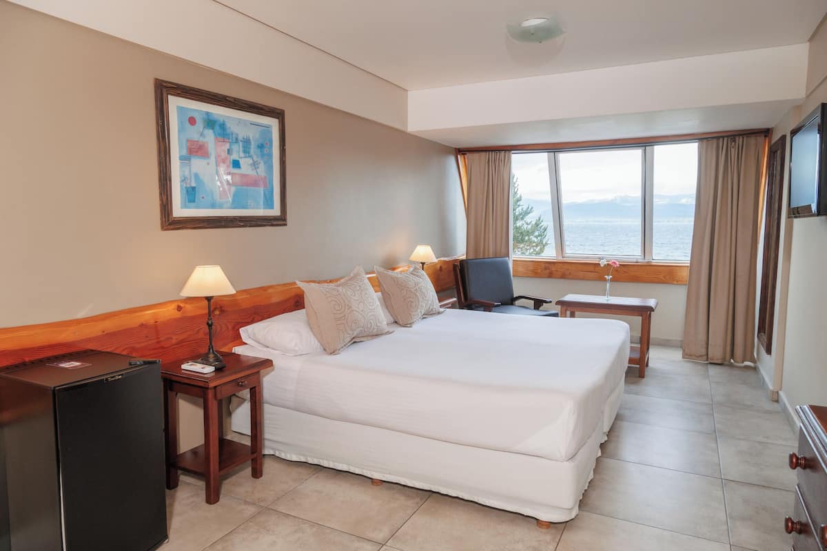 Habitación de hotel con una cama grande, un escritorio, una silla, una lámpara, un refrigerador y una ventana con vista al mar.