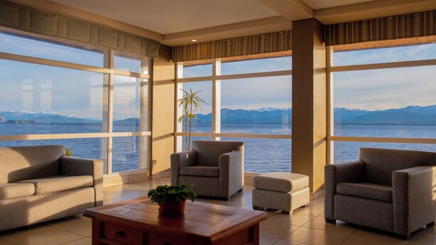 A spacious room with large windows offering a view of a body of water and mountains. There are two sofas and a coffee table with a plant. - Patagonia Hotel (San Carlos de Bariloche)