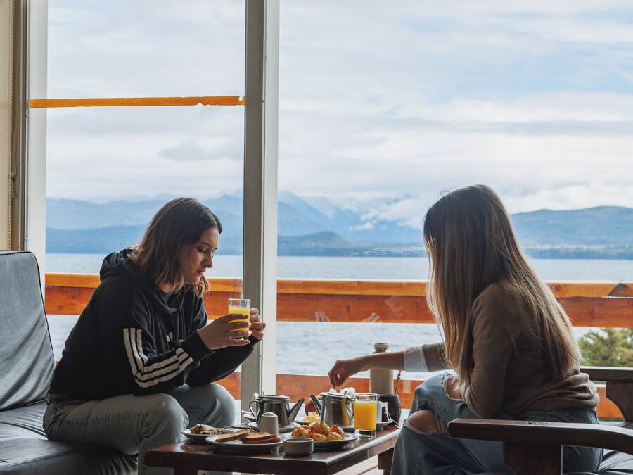 Twee personen zitten aan een houten tafel bij een groot raam met uitzicht op een wateroppervlak en bergen.