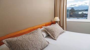 A hotel room with a bed, two pillows, a nightstand with a lamp, and a window with curtains.