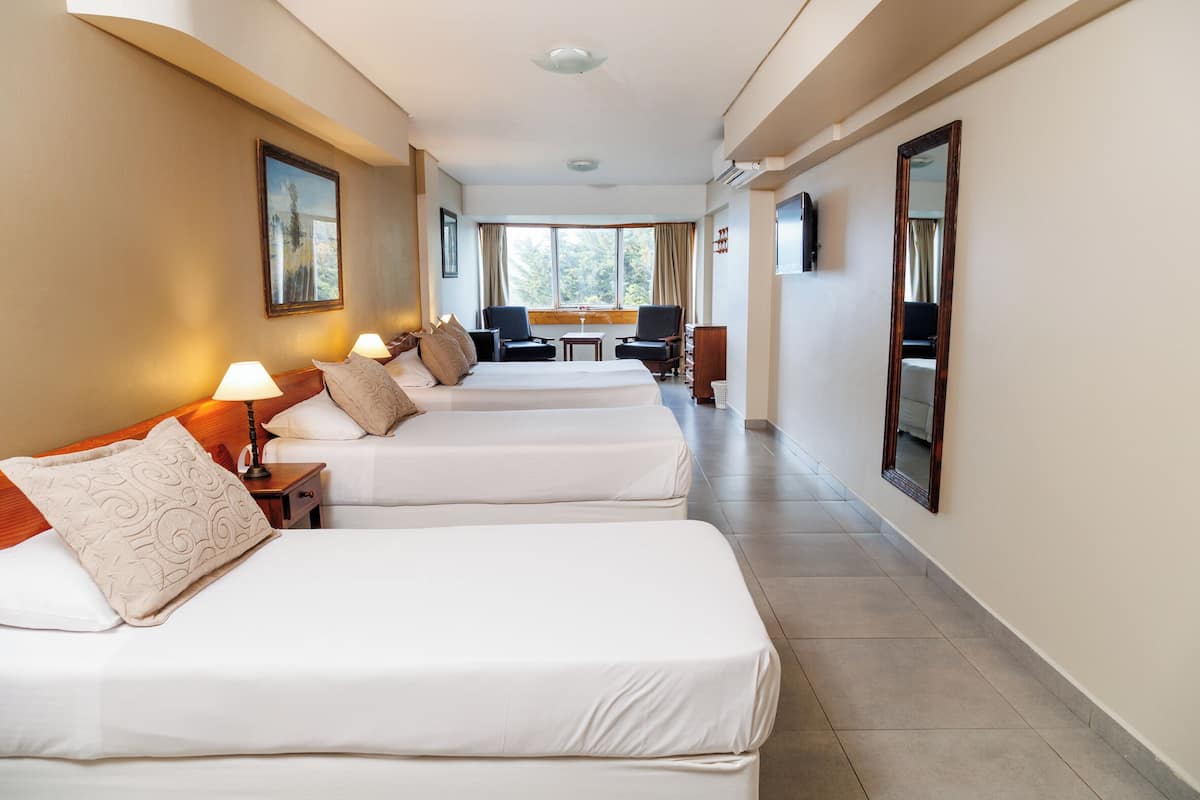 Habitación de hotel con dos camas, una mesita, sillas y un ventanal con vistas a árboles.