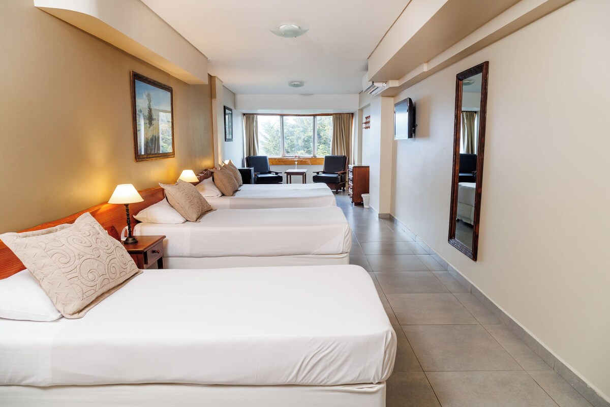 Habitación de hotel con dos camas, una mesita, sillas y un ventanal con vistas a árboles.