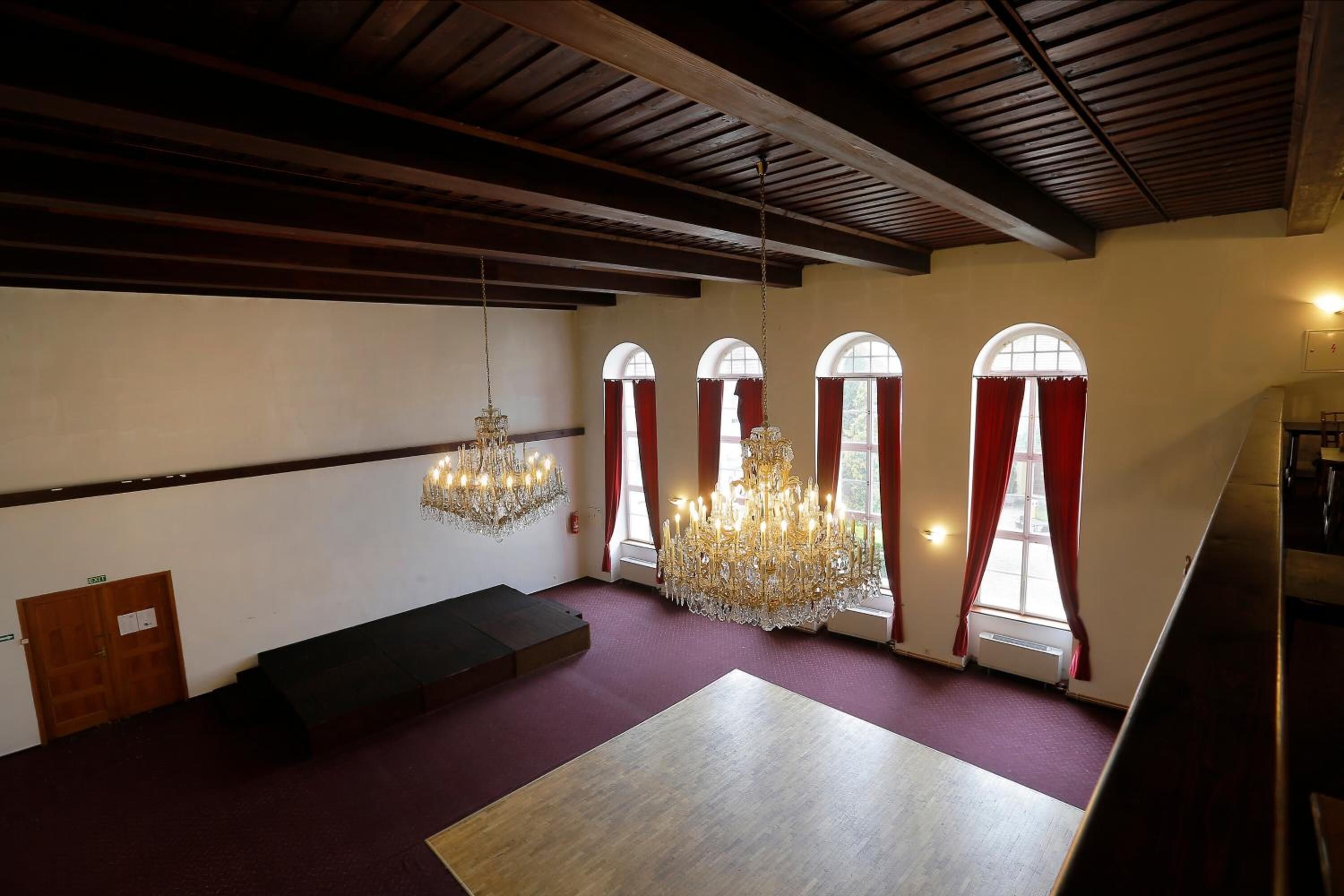 Una habitación amplia con techo alto, grandes candelabros, ventanas arqueadas con cortinas rojas y un banco de madera.