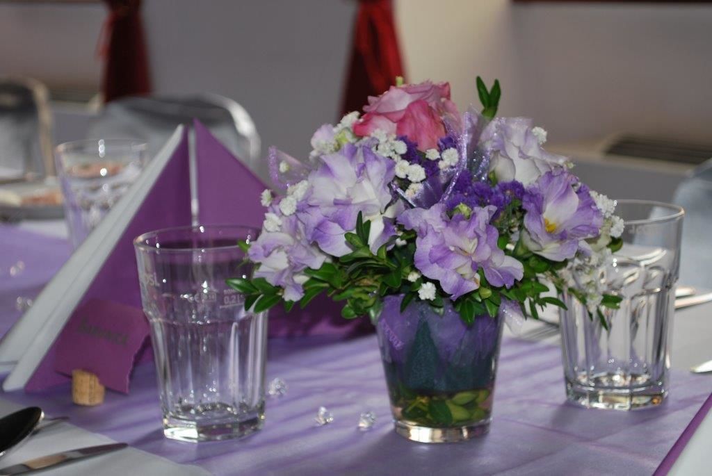 Une table recouverte d’une nappe violette, un vase en verre contenant des fleurs violettes et blanches, et deux verres vides.
