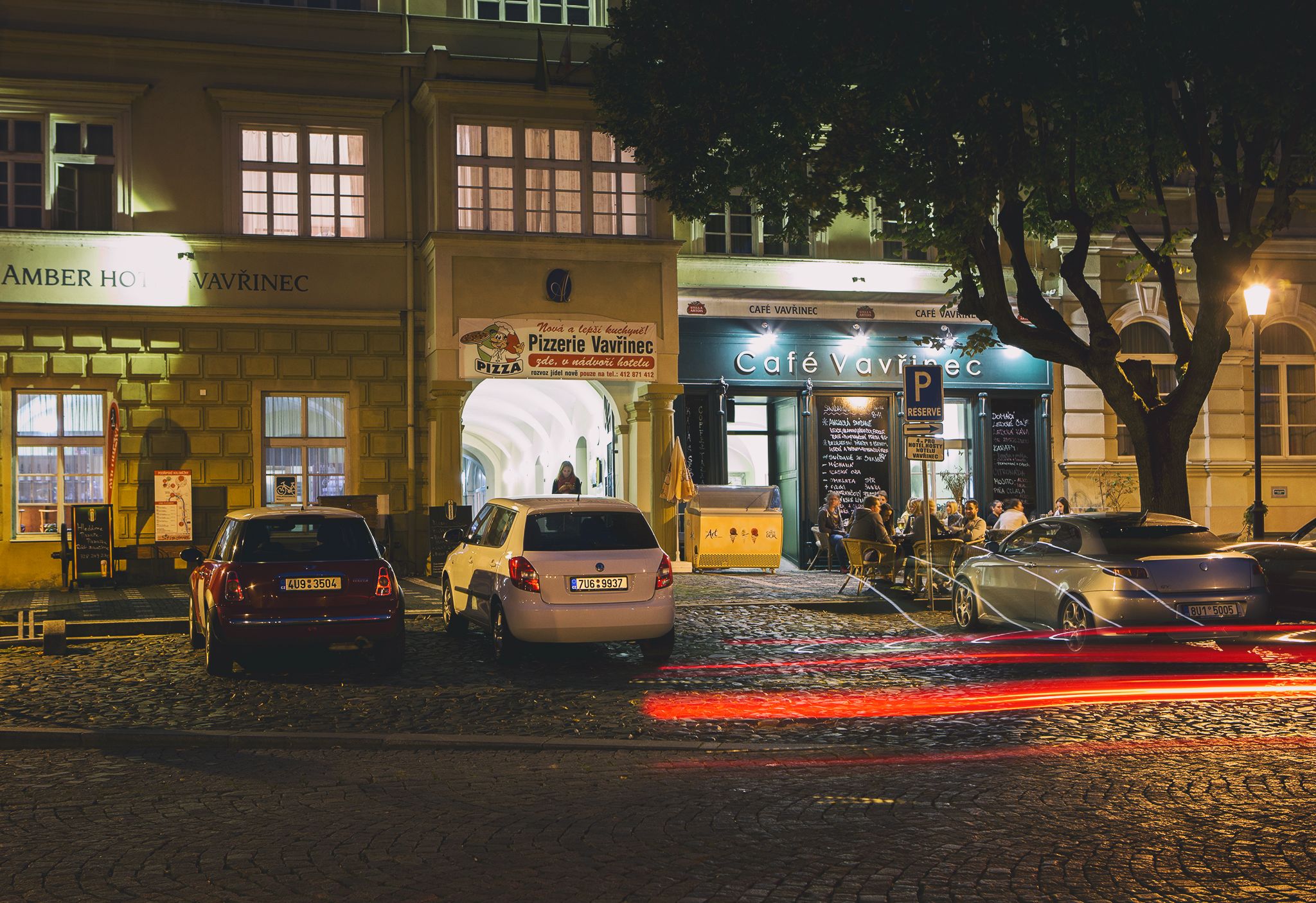 A cobblestone street with parked cars and a café named Café Vavrincé.